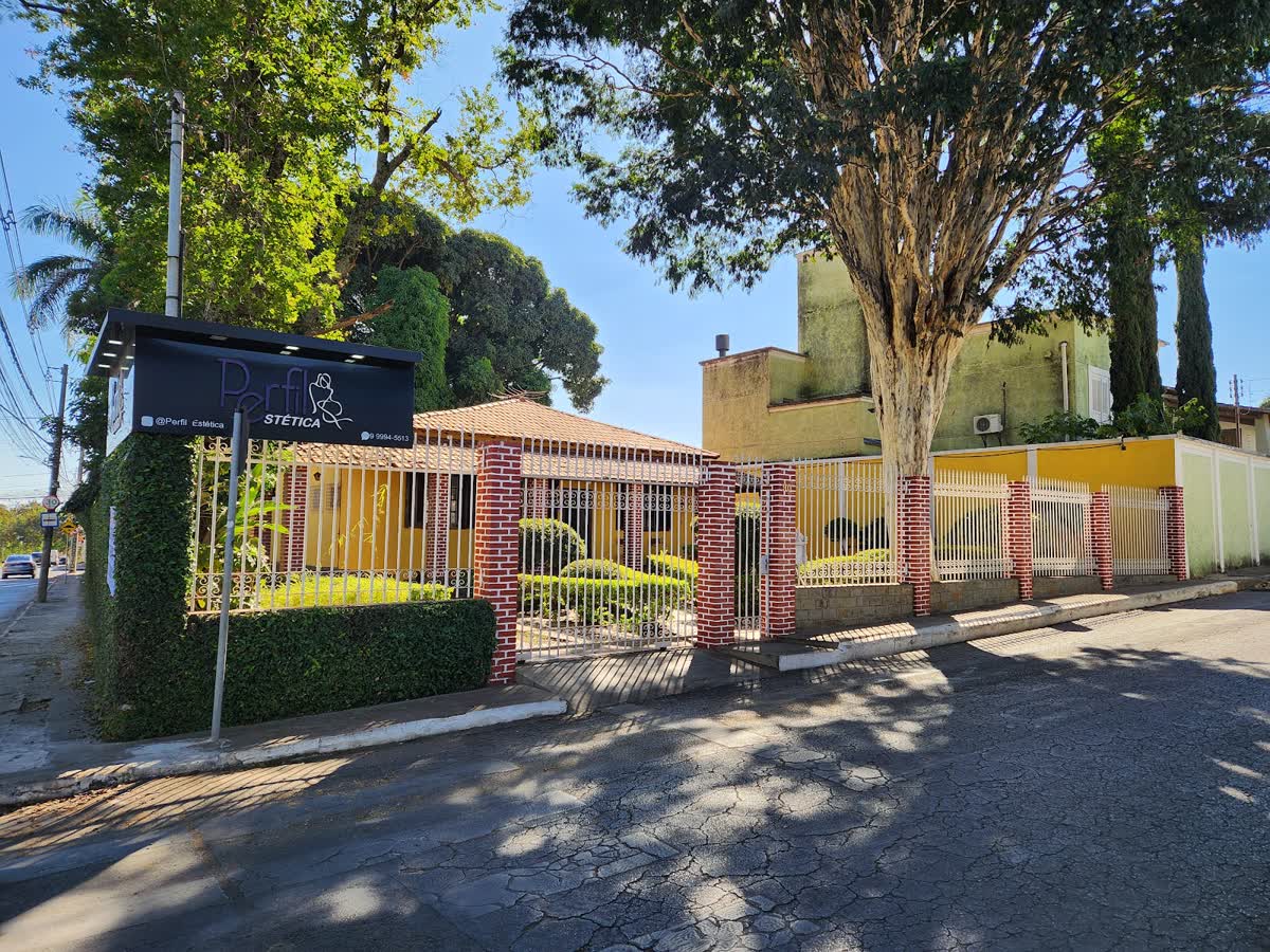 Fachada da Perfil Estética: a placa preta da clínica na esquina da casa amarela com jardim, no Três Barras